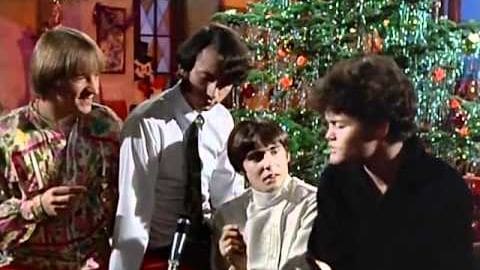 A group of young adults gathered indoors during Christmas, surrounded by festive decorations including a decorated tree and holiday-themed wall items.