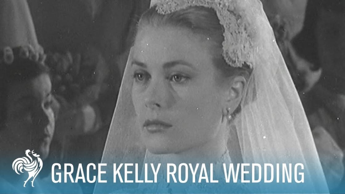 A bride at her royal wedding in Monaco, wearing a long white veil with lace trim and floral accents, pearl drop earrings, and a dark-colored suit with a tie or cravat. The setting appears to be indoors within a church or formal hall.