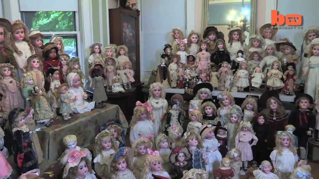 Indoor space filled with dolls displayed on tables under natural light from a window; various shades of beige, cream, and pastel tones dominate the scene.