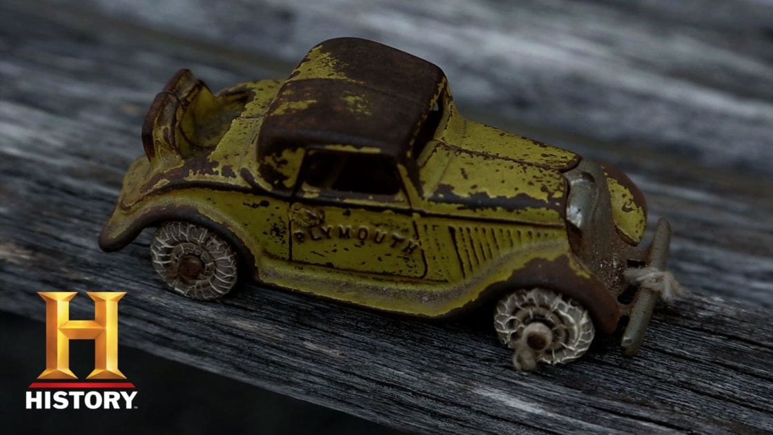 A small, vintage-style toy car labeled 'PLYMOUTH' sits atop a rustic wooden surface outdoors under natural light.