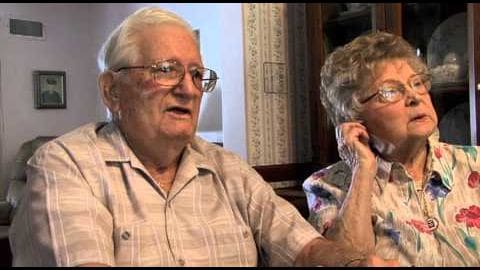 An older adult male and female couple seated indoors, wearing striped button-up shirt and floral-patterned blouse respectively, with soft, even lighting suggesting daytime.