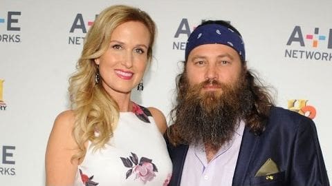 An adult female in a white floral dress with black accents and large hoop earrings at an A+E Networks event, alongside an adult male in a dark blue blazer over a light-colored shirt with a pocket square and navy bandana on his head.