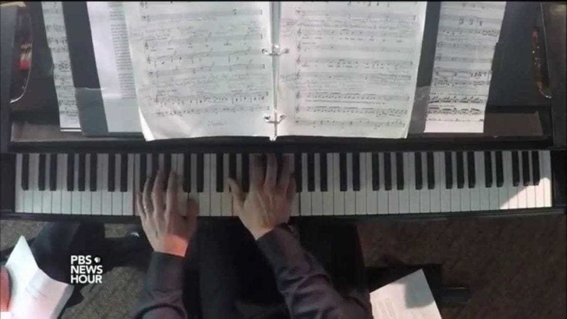 Adult male pianist playing on a piano with sheet music, PBS NewsHour logo visible, indoor setting with carpeted floor.
