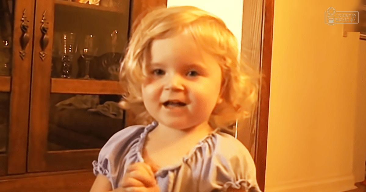 A toddler in a light blue dress indoors, surrounded by wooden furniture and home decor.