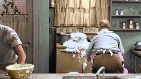A medium-sized dog with light brown fur and darker markings, wearing no collar, stands by a kitchen counter where two individuals are engaged in activities; wooden cabinets, checkered curtains, and various kitchen items create the setting.