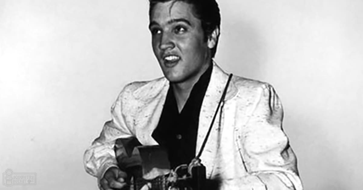 Young adult male wearing a white puffed-sleeve jacket over a fitted black v-neck shirt, holding a guitar indoors with plain white walls as background.