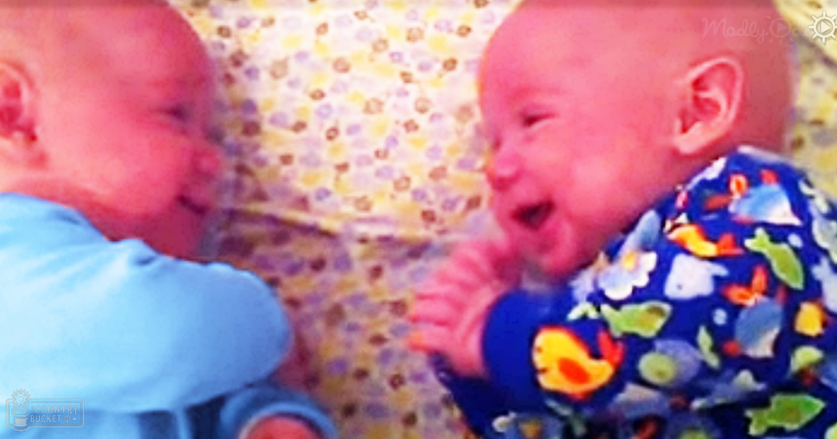 Two infants wearing blue garments with colorful patterns and a floral-patterned backdrop, suggesting an indoor home setting.