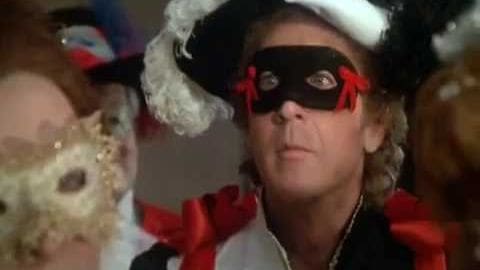 An older adult male in a festive setting with curly hair wearing a white hat adorned with black and red decorations at a masquerade event, surrounded by others dressed similarly.