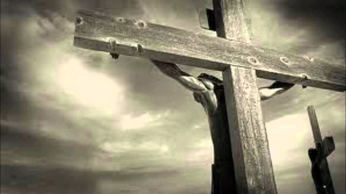 Two figures on wooden crosses against a dramatic, somber sky in an outdoor setting under muted sepia-toned light.