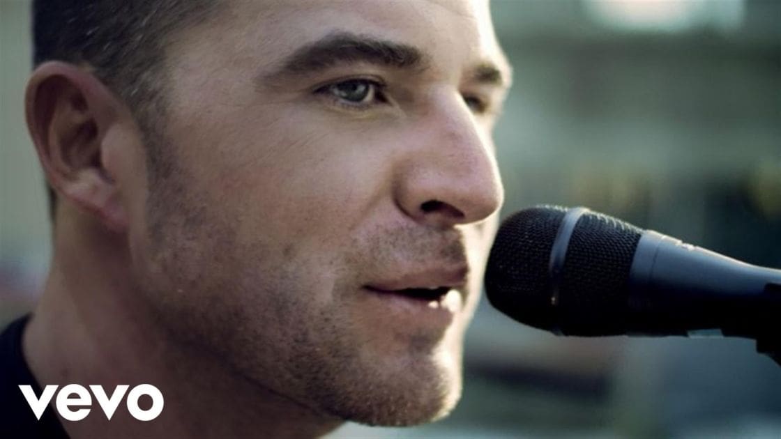 A middle-aged adult male singing into a microphone in an outdoor setting during daylight hours.