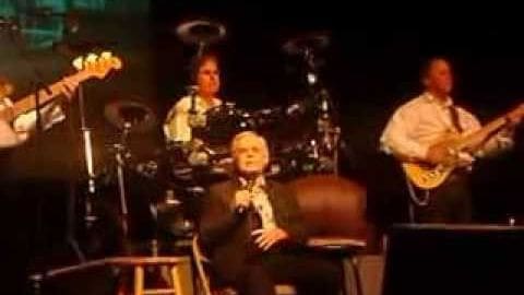 Child male performer in school uniform sits on stage with adult male drummer and guitarist; dog stands nearby under dim indoor lighting.
