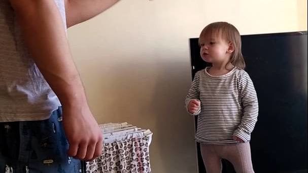 A toddler female in a striped shirt stands next to an adult male wearing jeans with graphic print indoors under natural light.