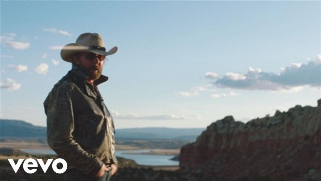 Adult male in a desert landscape, wearing a camouflage-patterned western-style jacket with a high collar and long sleeves, wide-brimmed cowboy hat, and sunglasses. The scene features rocky formations, a body of water under clear skies.