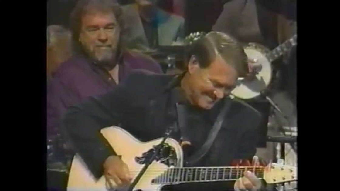 An adult male musician playing an electric guitar on stage, wearing a dark shirt and accessories for the instrument; another middle-aged to older adult male in the background wearing a purple long-sleeve shirt.