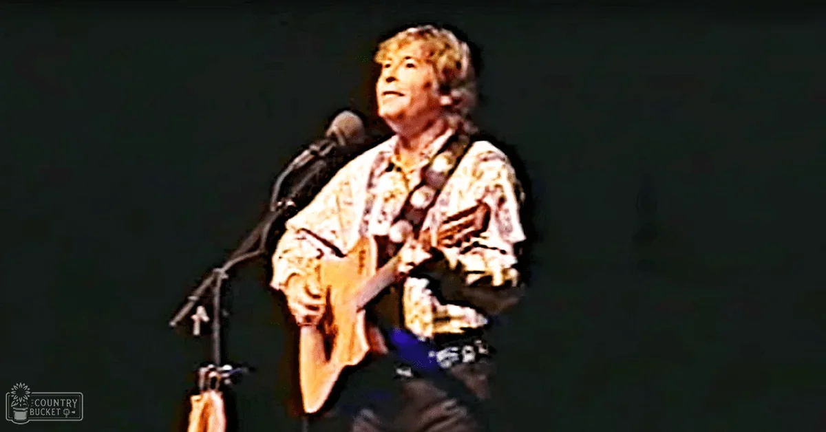 A middle-aged male performer on stage wearing a patterned shirt with earthy tones and dark pants, holding a guitar strap while performing at an indoor concert venue under focused stage lighting.