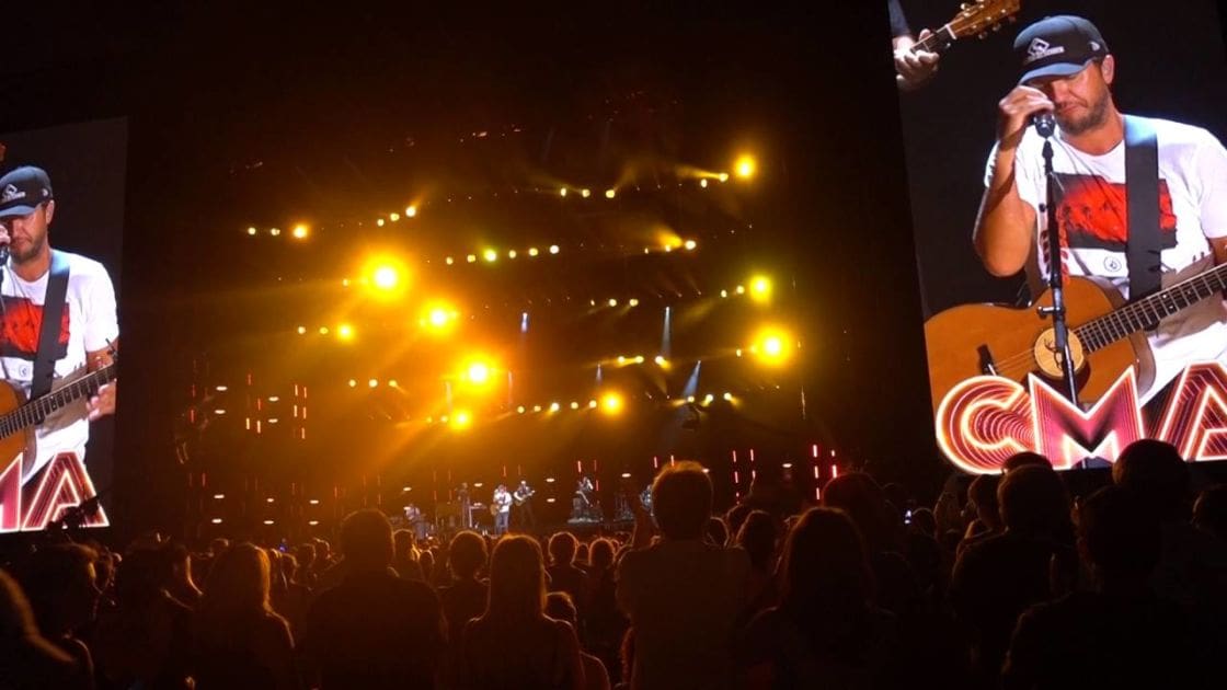 A country music concert at the CMA Awards takes place in an indoor arena, with bright yellow stage lights and a large screen displaying the performer.