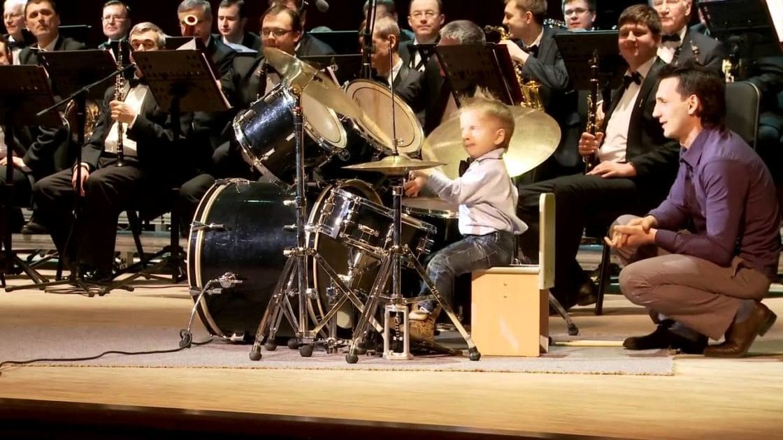 A young child plays drums in front of an orchestra at what appears to be a concert hall. The scene is filled with adult musicians seated and playing various instruments.