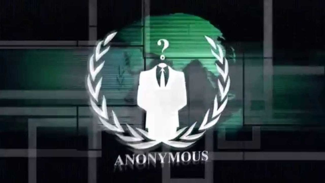 A nighttime urban scene with 'Anonymous' logo against dark glass windows reflecting green light.