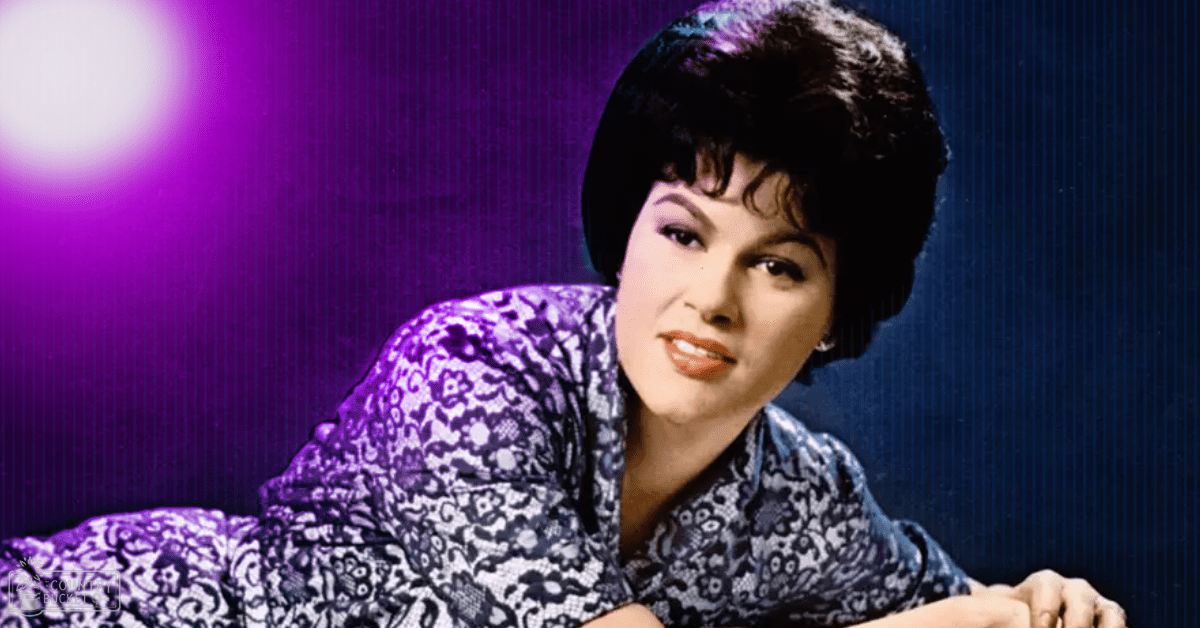 An adult female wearing a loose, mid-20th-century blue and white patterned blouse in an indoor setting with a dark blue and purple gradient background.