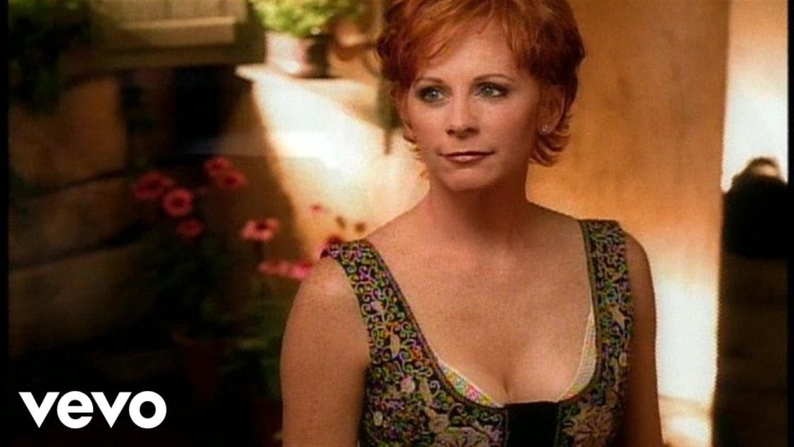 A middle-aged adult female wearing a sleeveless floral dress outdoors with potted plants and a stone bench in the background.