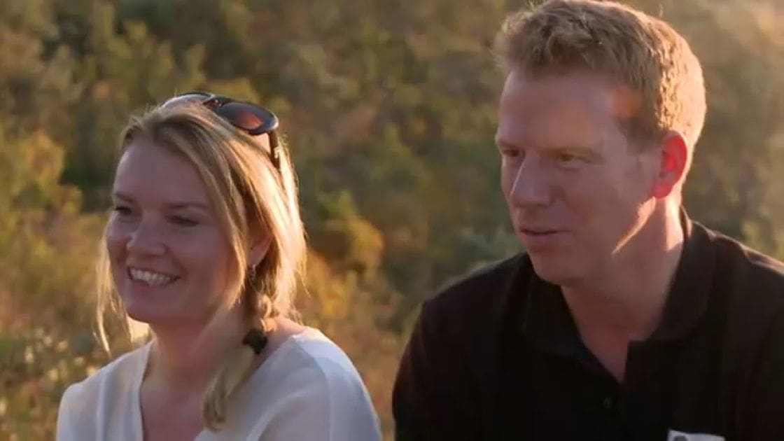 An adult male singer wearing a black polo shirt with short sleeves, likely made of cotton or polyester blend, alongside an adult female in a white blouse or top with a relaxed fit and short sleeves, set against an outdoor backdrop with trees and foliage in golden hues suggesting late summer or early autumn, bathed in warm tones from sunrise or sunset.