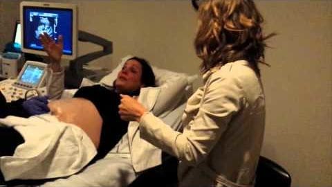 An adult female medical professional wearing a white lab coat and an adult female patient lying down in a medical examination room, with medical ultrasound equipment displaying images of a fetus.