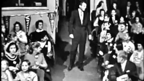 A man in a dark suit walks down a runway or stage at an indoor event with seated audiences.