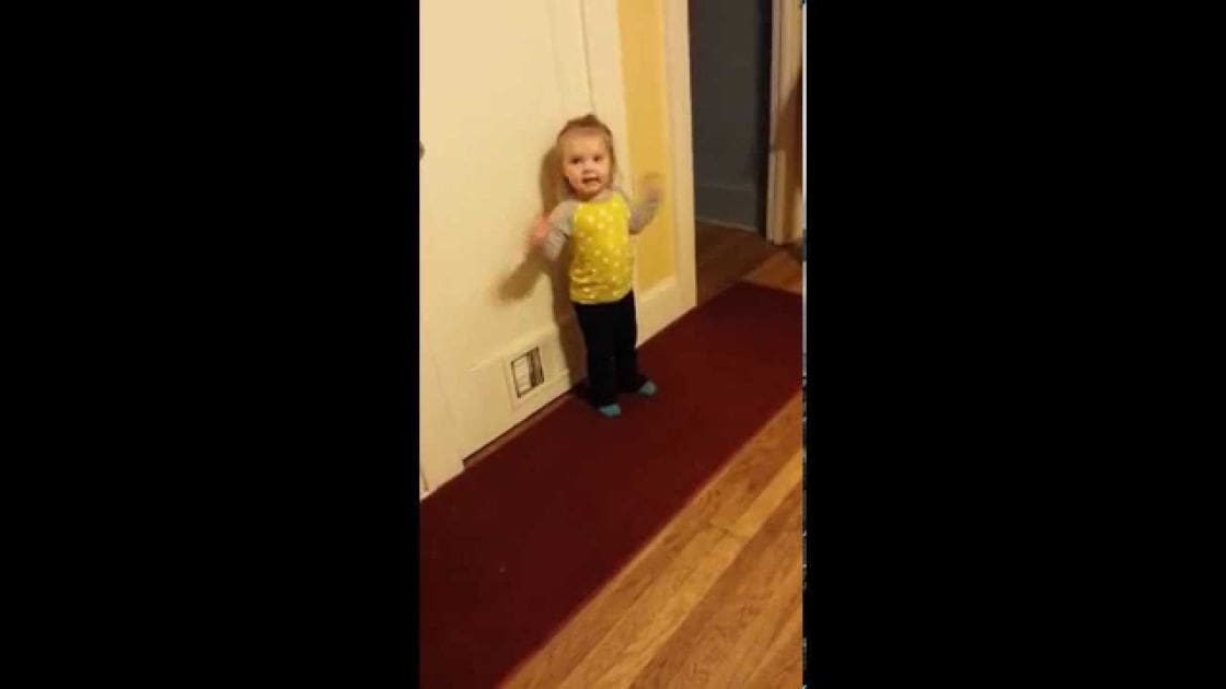 A toddler in a yellow polka dot shirt and black pants stands indoors with artificial lighting, surrounded by carpeted floors and indoor furniture.