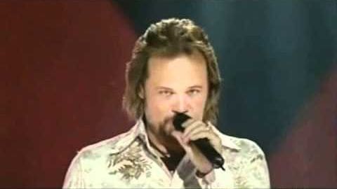 An adult male singer in a light-colored, patterned floral shirt with long sleeves and collar, performing on stage holding a microphone against a backdrop of deep red and darker blue curtains or stage decorations.