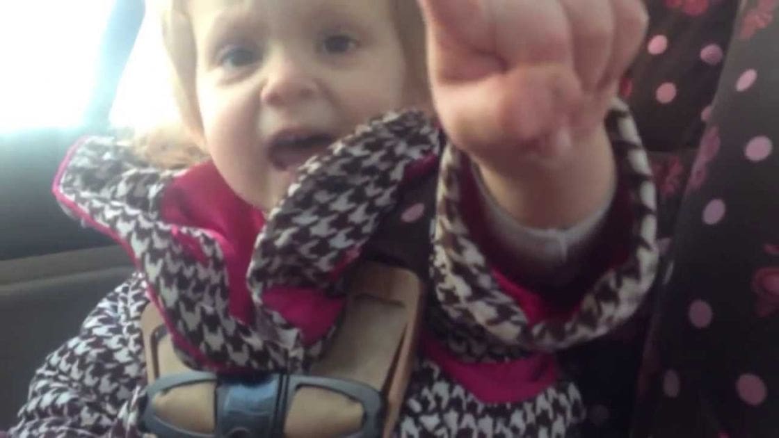 An infant/toddler girl wearing a patterned scarf with black, white, pink, and brown colors sits in a car seat inside a vehicle. The setting is bright daylight, possibly spring or summer.