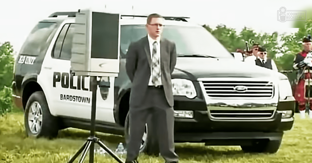 An adult male in a dark gray suit with patterned tie stands before a police SUV labeled 'POLICE BARDSTOWN' amidst an outdoor setting with grassy terrain, trees, and a bagpipe player in traditional attire.