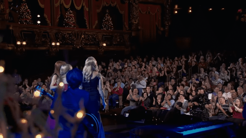 Performers on stage wearing blue dresses with high-heeled shoes; audience members in casual or semi-formal attire with accessories;