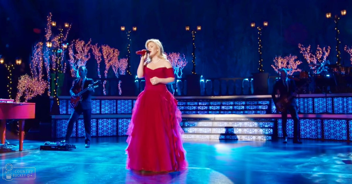 A woman performing on stage at an indoor concert venue with a red off-the-shoulder gown and ruffled layers, surrounded by musicians in dark suits under artificial lighting.