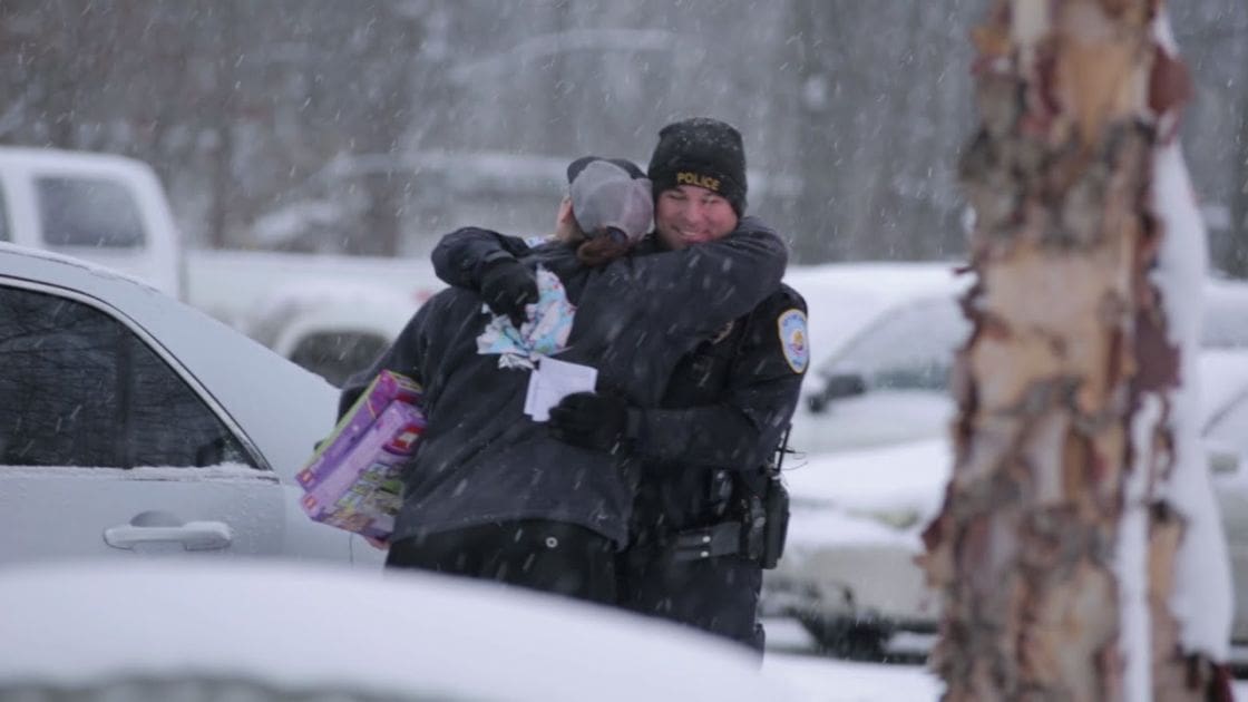 An adult male police officer hugging an adult female in a snowy community setting with snowflakes falling, surrounded by trees and vehicles covered in snow