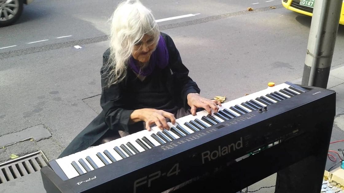 An older adult female street performer wearing a black coat with purple scarf and black pants, playing an electronic keyboard in an urban outdoor setting with asphalt, drain, yellow vehicle visible, likely city or town center, natural daylight with shadows indicating daytime.