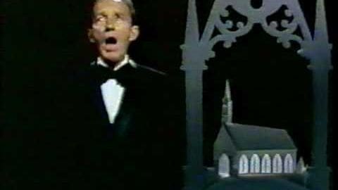 A young adult male in a fitted tuxedo sings against a church backdrop, likely an indoor performance or recording.