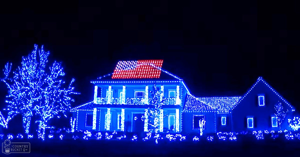 A festive outdoor residential setting with a house decorated with blue and white Christmas lights, red and white lights arranged to resemble an American flag on the roof, and additional decorative lights around the base. The scene is dark, suggesting evening or night time.
