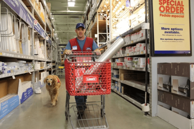 An adult male wearing a blue t-shirt under a red Lowe's vest, pushing a shopping cart in a hardware store with a golden retriever dog nearby.