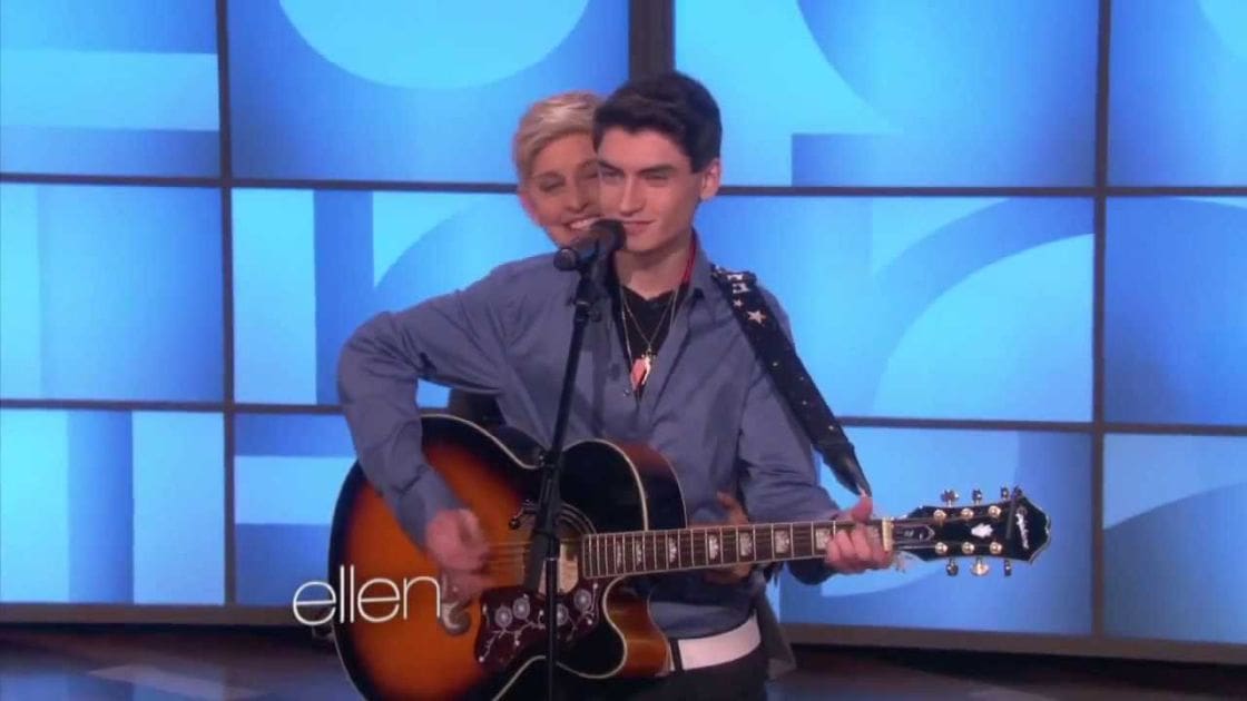 A young adult male playing a guitar on 'The Ellen Show', wearing a gray button-up shirt, dark pants or jeans, and a necklace with a pendant.