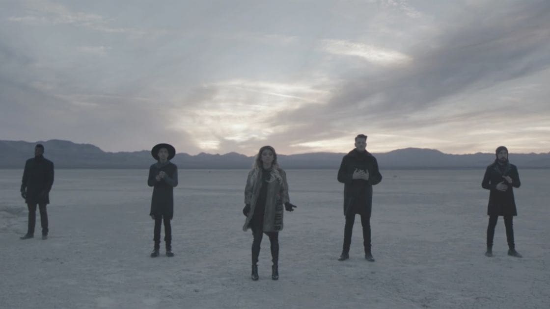Five adults standing together in a vast open landscape under a cloudy sky; muted tones of gray, blue, white, and beige dominate the scene.