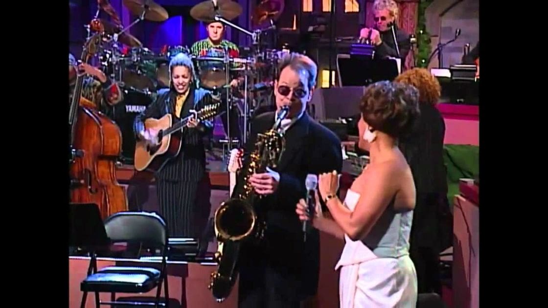 An adult male singer holding a saxophone on stage with an adult female in a sleeveless dress, surrounded by musical instruments like drums and guitars under artificial stage lights.
