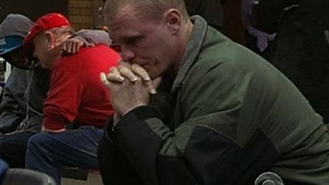 Young adult male wearing a dark green jacket with black sleeves; older adults in casual attire including red shirts, blue jeans, gray hoodies, and backward caps; urban setting with natural daylight suggesting cool to cold weather.