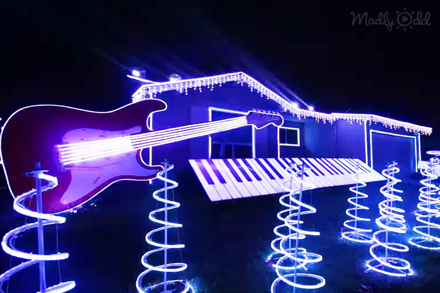 An outdoor nighttime display with illuminated musical instruments like a guitar and piano, surrounded by spiral lights, with 'Madly Odd' text visible in the top right corner.