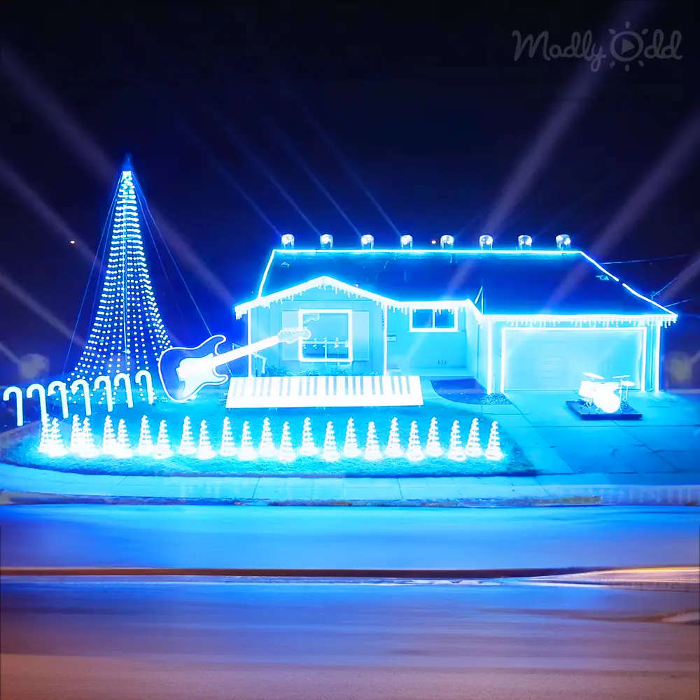 A residential street at night adorned with Christmas lights, with a lit-up electric guitar, keyboard, and drum set, creating a festive winter atmosphere.