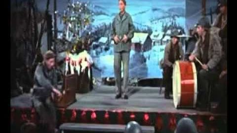 A group of adult males in a performance setting on an indoor stage with a snowy landscape backdrop and a large red drum, suggesting a musical or theatrical production during winter.