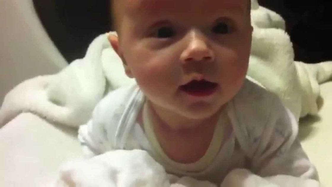 An infant wearing a white, patterned onesie indoors with warm indoor lighting and a dark background.