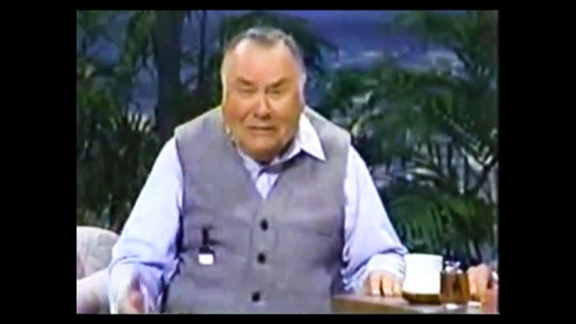An older adult male in a studio setting with light blue button-up shirt and grayish-blue vest, surrounded by greenery and artificial lighting.