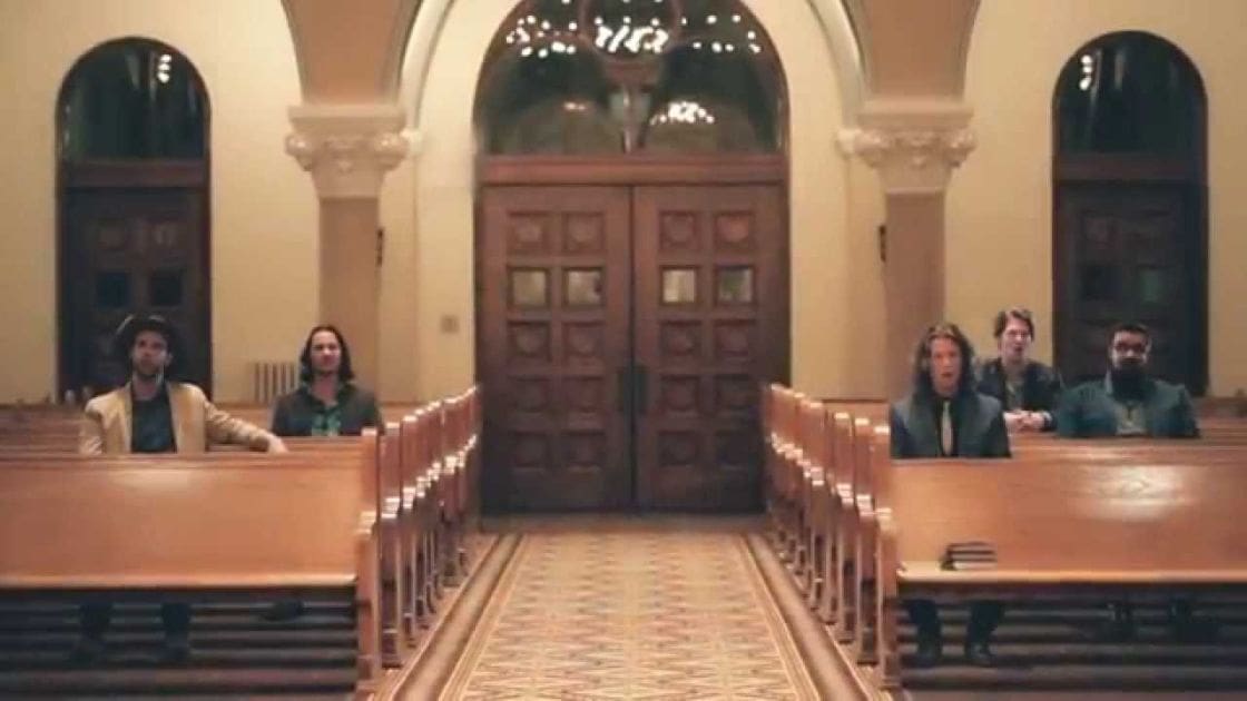 A group of individuals indoors resembling a church or cathedral setting with wooden pews, arched windows, and ornate double doors.