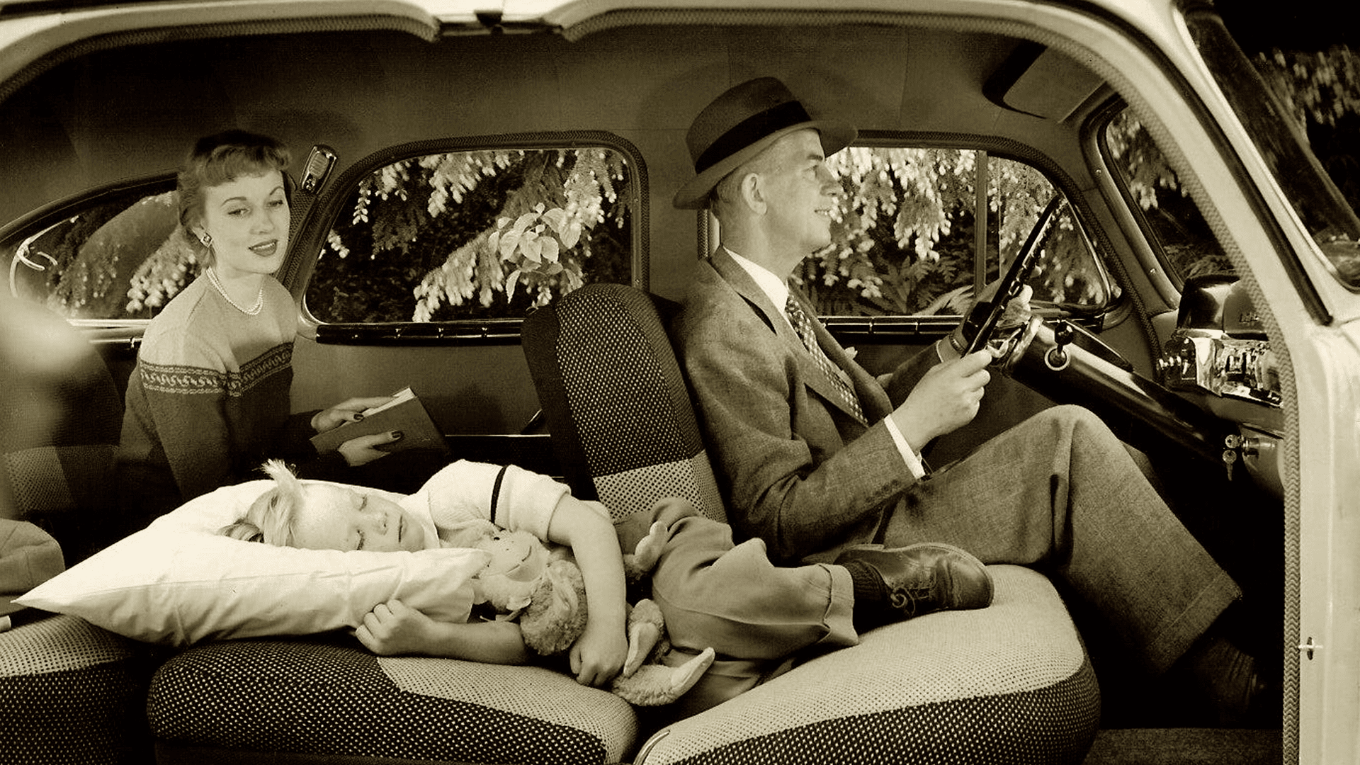 A family inside a vintage car, likely mid-20th century, with an adult female in a patterned sweater and pearl necklace, an adult male in a suit holding what appears to be a shotgun, and a child sleeping on a pillow in pajamas. The setting includes a suburban or rural landscape with trees outside the car windows.