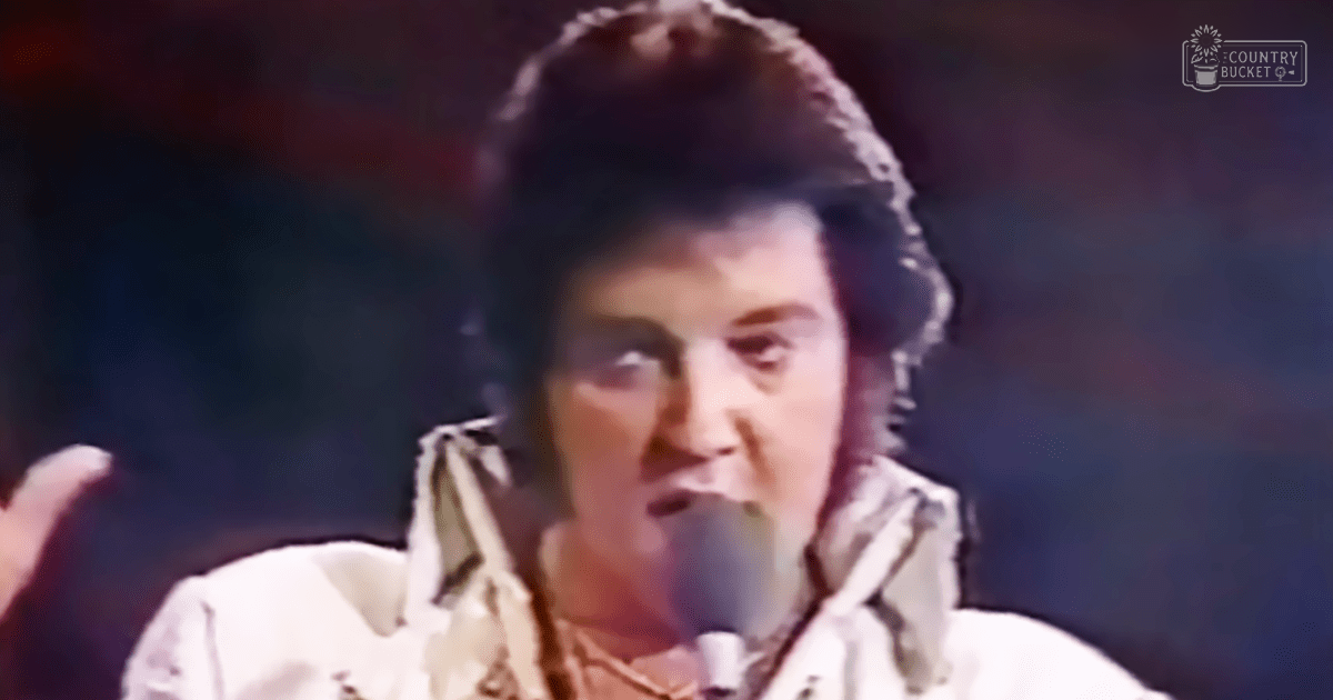 An adult male singer wearing a white collared shirt from an earlier era sings into a microphone on a dimly lit stage at what appears to be a concert or performance venue.